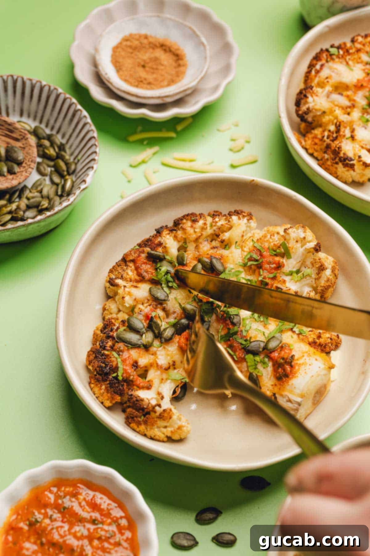fork and knife cutting into a cheesy cauliflower steak with pepitas and salsa on top. 