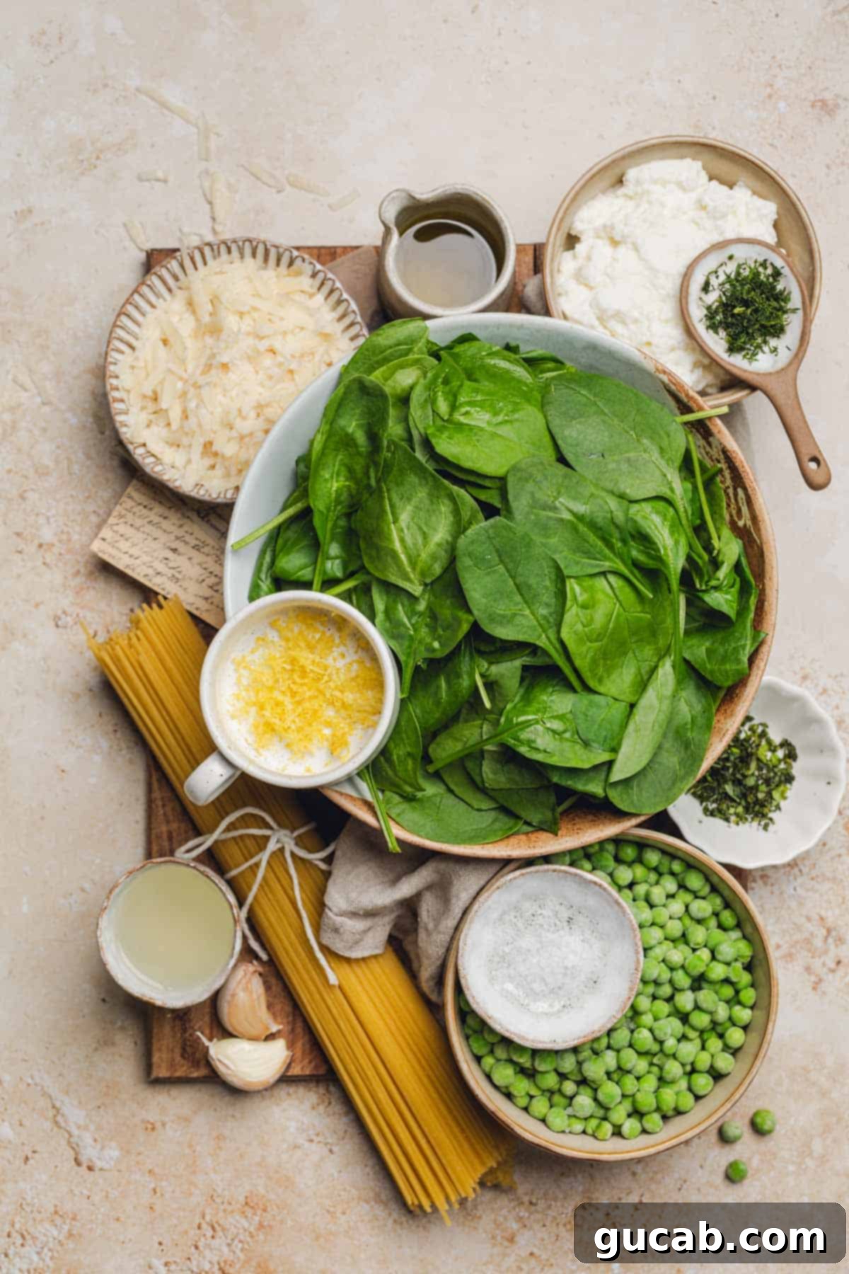 A collection of fresh ingredients laid out, including a bowl of spinach, lemon zest, parmesan, ricotta cheese, fresh dill, peas, lemon juice, minced garlic, and uncooked spaghetti noodles.