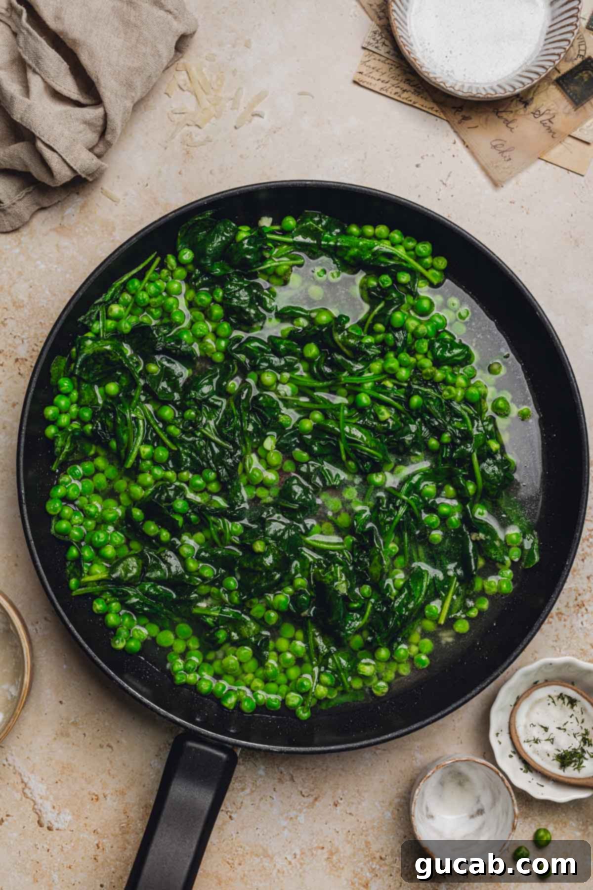 A skillet showing cooked spinach, peas, and the addition of pasta water, forming the base for the creamy sauce.