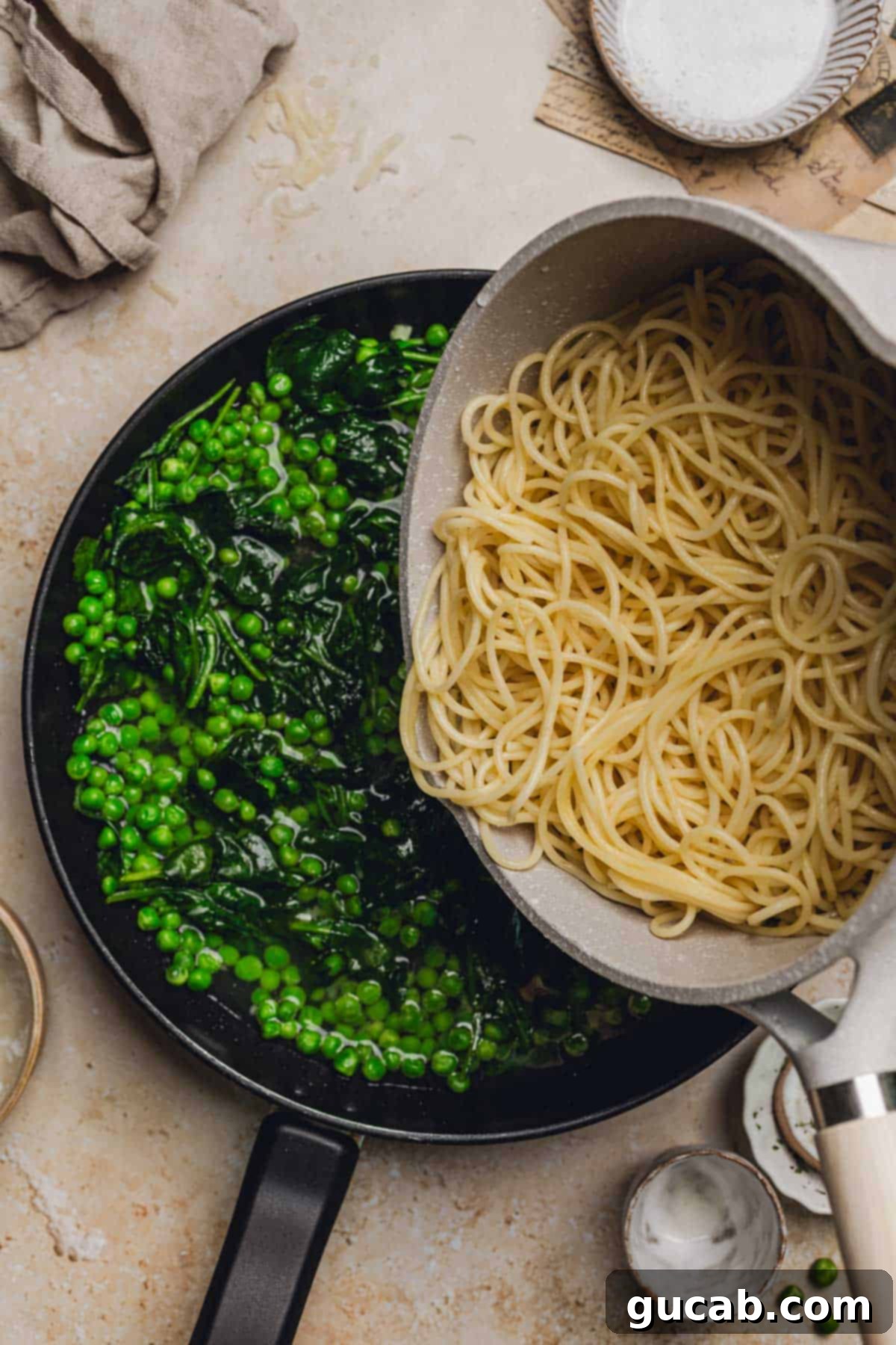 A hand pouring the cooked spaghetti pasta into the skillet, joining the peas, spinach, and pasta water.