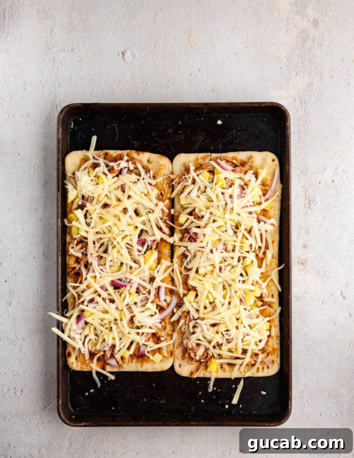 A flatbread fully topped with shredded chicken, pineapple, thinly sliced red onion, and generous amounts of shredded cheese, perfectly arranged for baking.