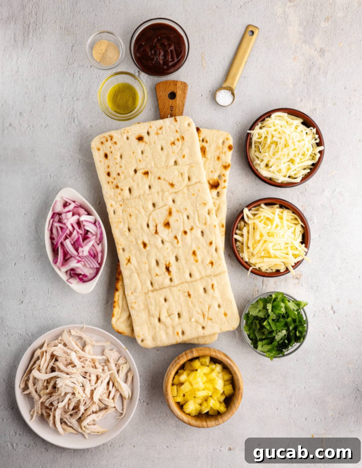 A collection of fresh ingredients for flatbread pizzas including thinly sliced red onion, a bottle of barbecue sauce, olive oil, a bowl of spices, blocks of gouda and mozzarella cheese, fresh cilantro, and a bowl of pulled chicken.