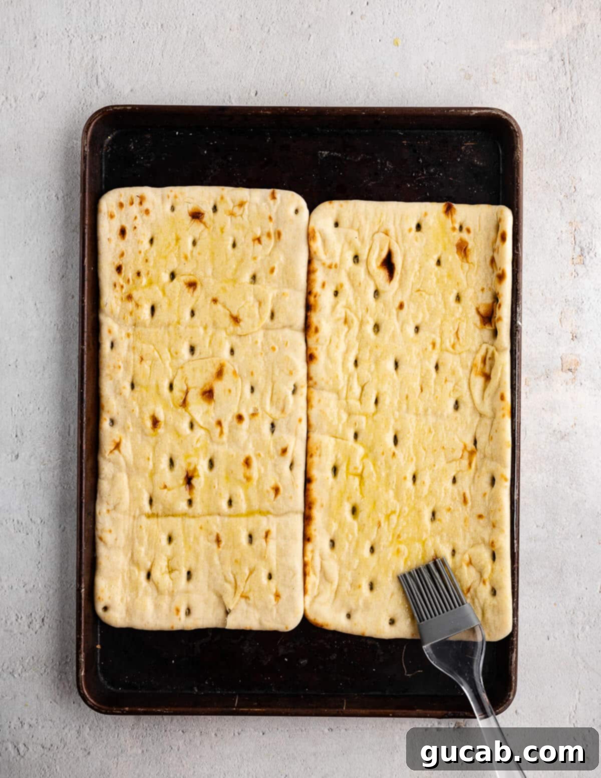 A hand brushing olive oil onto flatbreads arranged on a sheet pan, preparing them for seasoning.