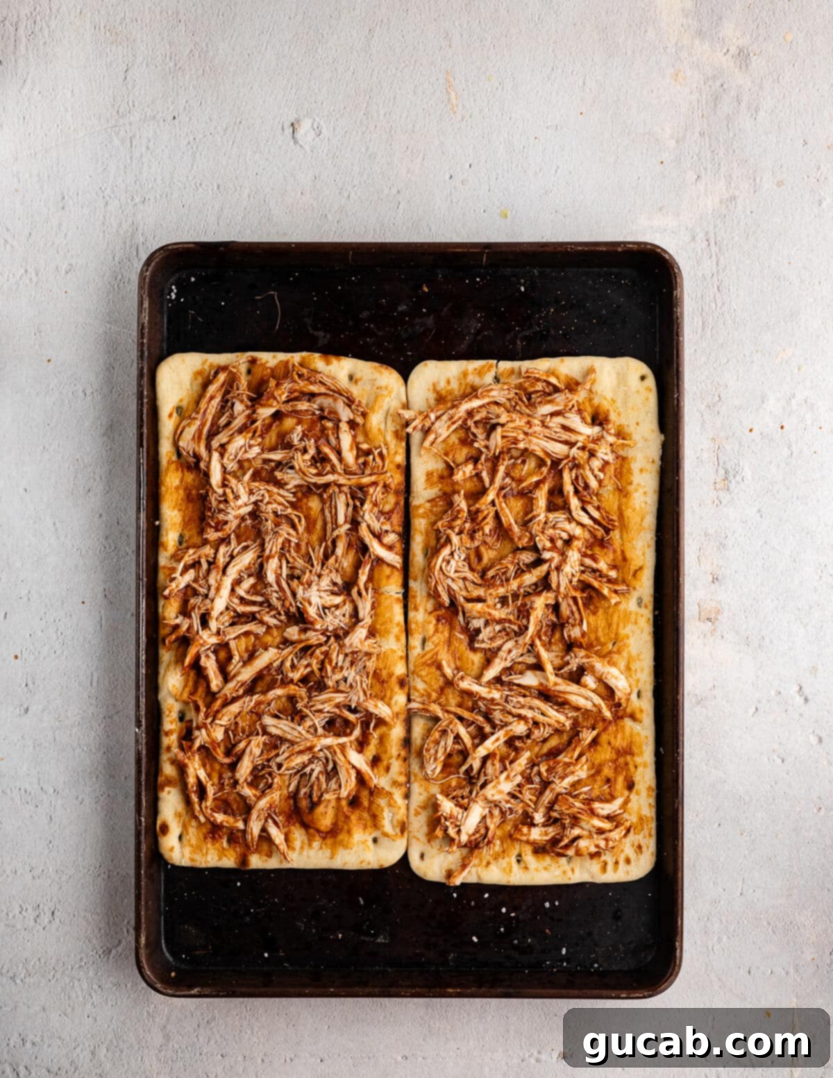 A flatbread coated in barbecue sauce, topped with shredded chicken, on a sheet pan, showing the even distribution of ingredients.