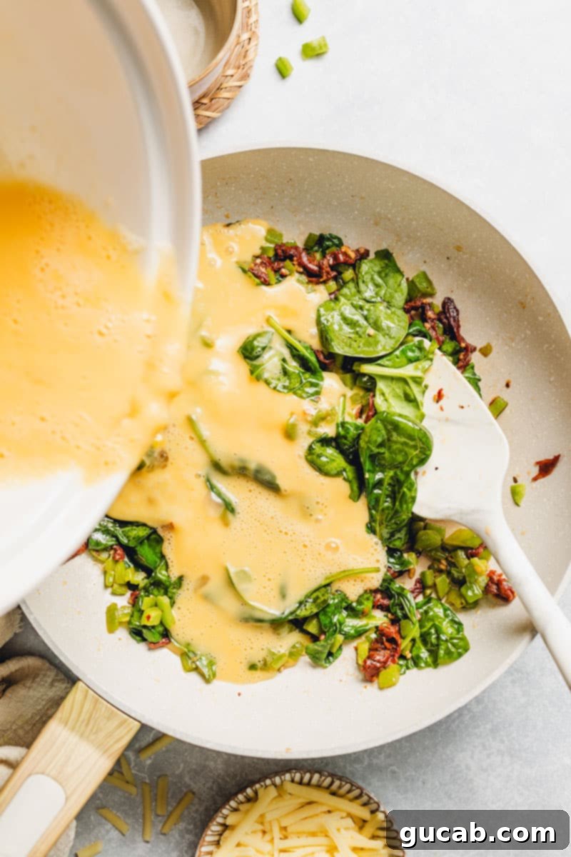 pouring eggs into a skillet with cooked spinach, bell pepper, and sundried tomatoes