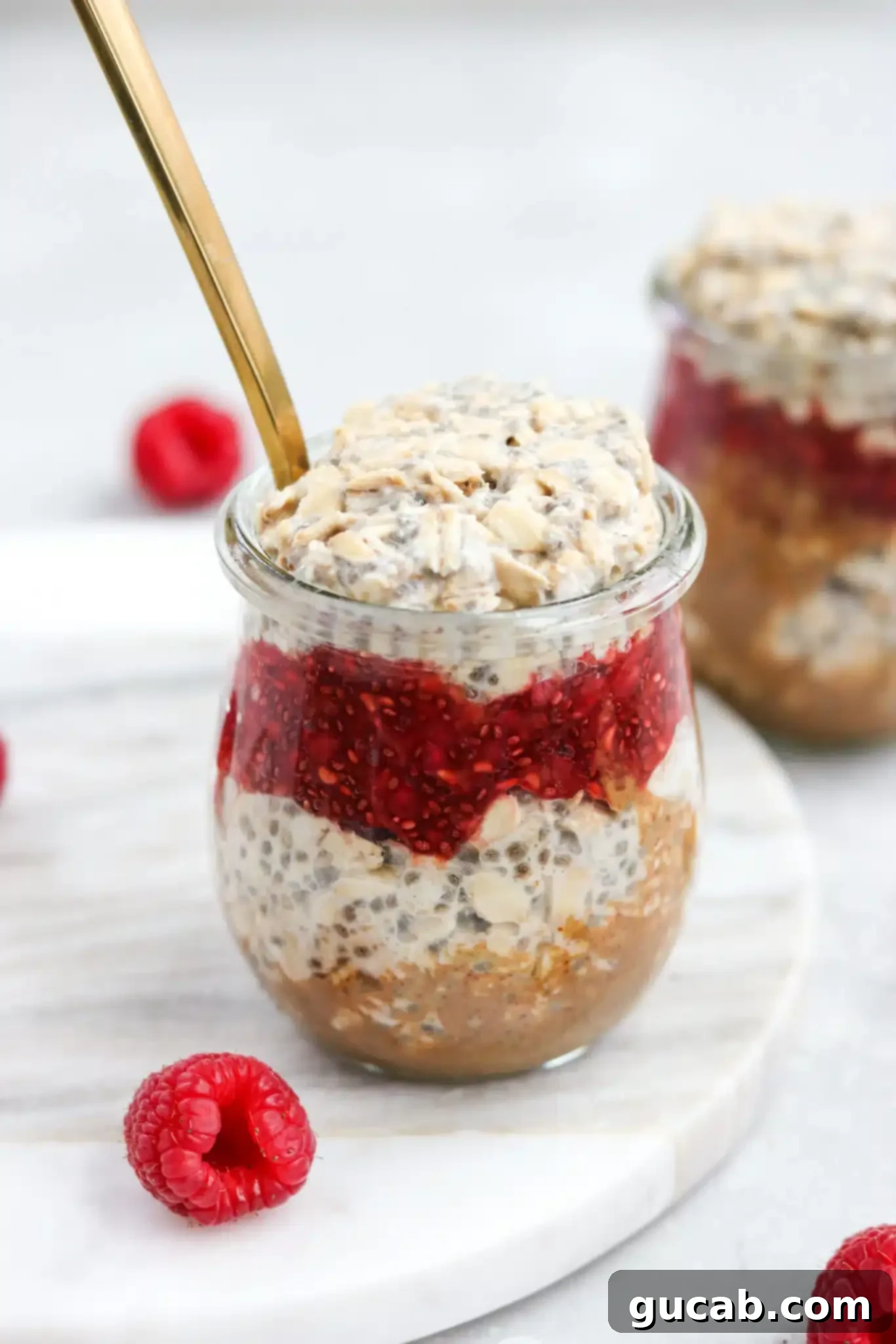 A charming jar layered with creamy almond butter, vibrant raspberry chia jam, and wholesome overnight oats, reminiscent of a classic PB&J sandwich.