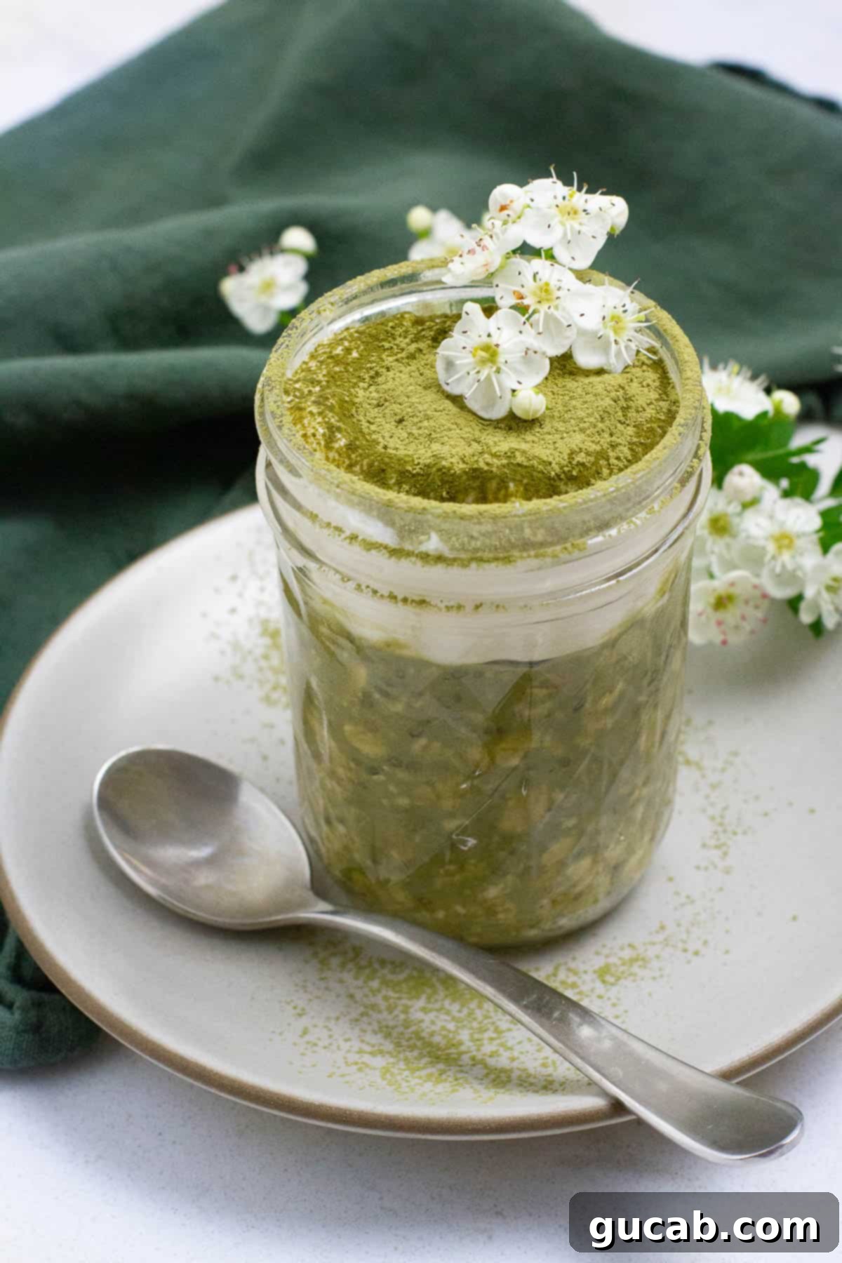 A glass jar of vibrant green matcha overnight oats, elegantly topped with a dusting of matcha powder and delicate white edible flowers, creating a serene and healthy breakfast.