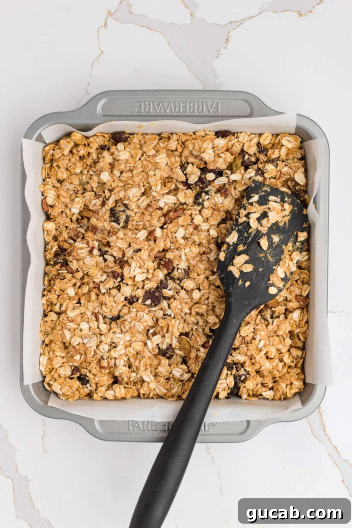 A spatula carefully smoothing out the soft granola mixture within a square baking dish, working towards an even and consistent layer.