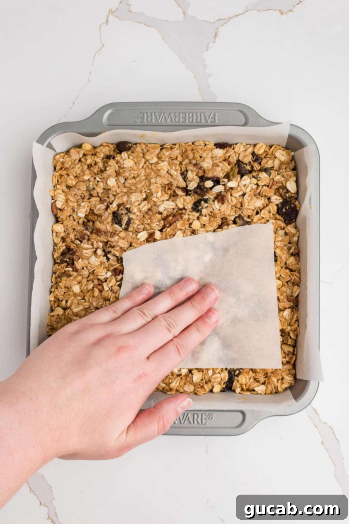 A hand using a piece of parchment paper to firmly press the granola mixture into the baking dish, compacting it tightly for stable and non-crumbly bars.