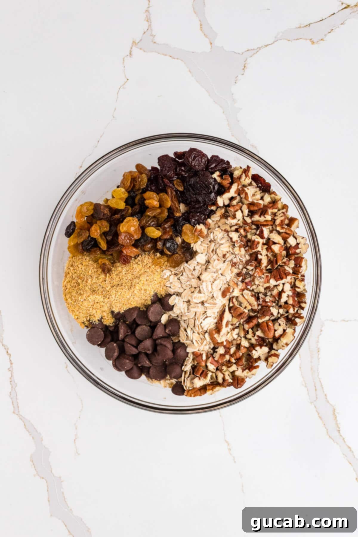 A large mixing bowl with the wet ingredients topped with a colorful array of golden raisins, tart dried cherries, crunchy chopped pecans, hearty oats, and nutritious golden flax meal, all waiting to be mixed into the granola bar batter.