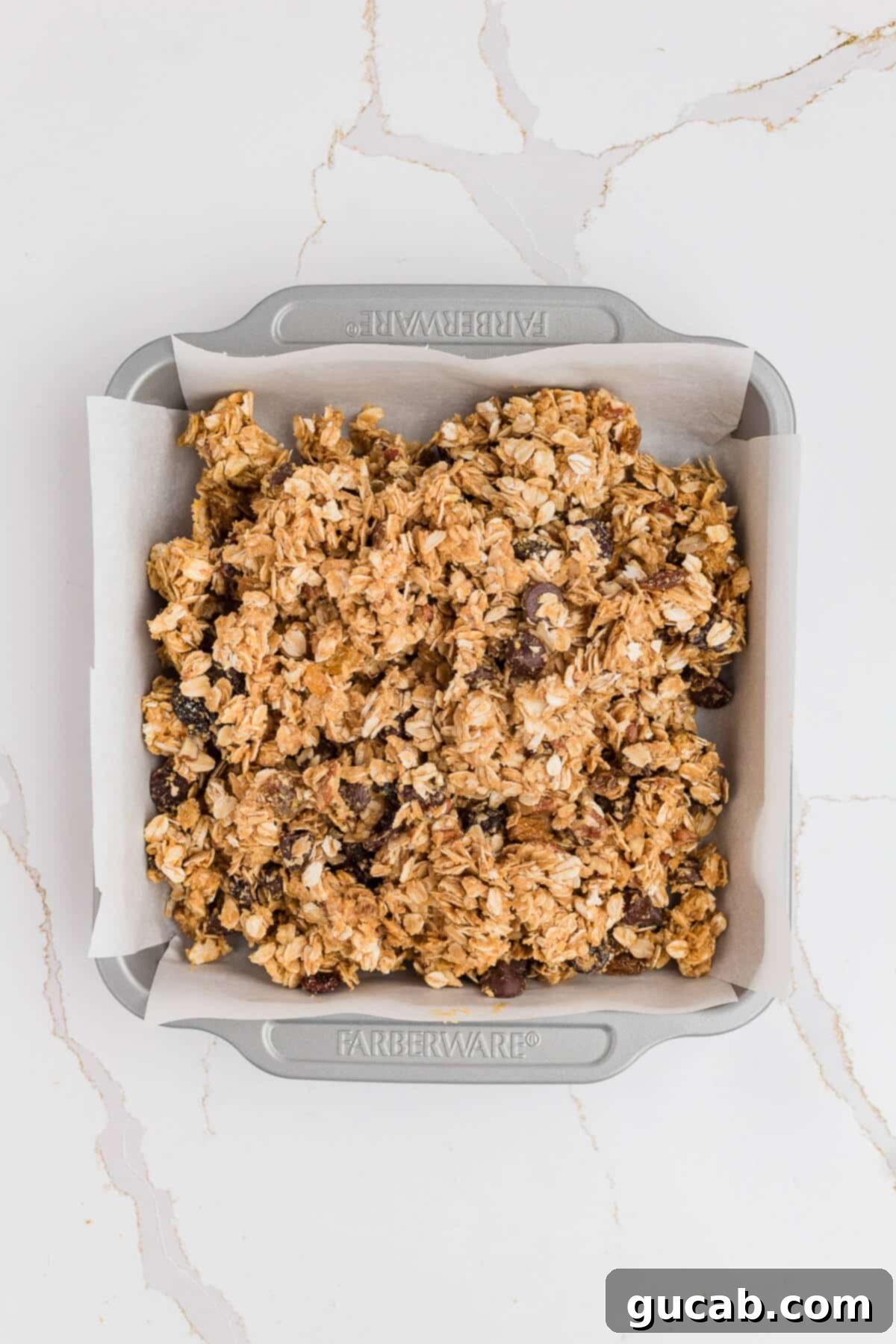 The granola bar mixture loosely transferred into the parchment-lined baking pan, spread somewhat evenly but still requiring firm compression.