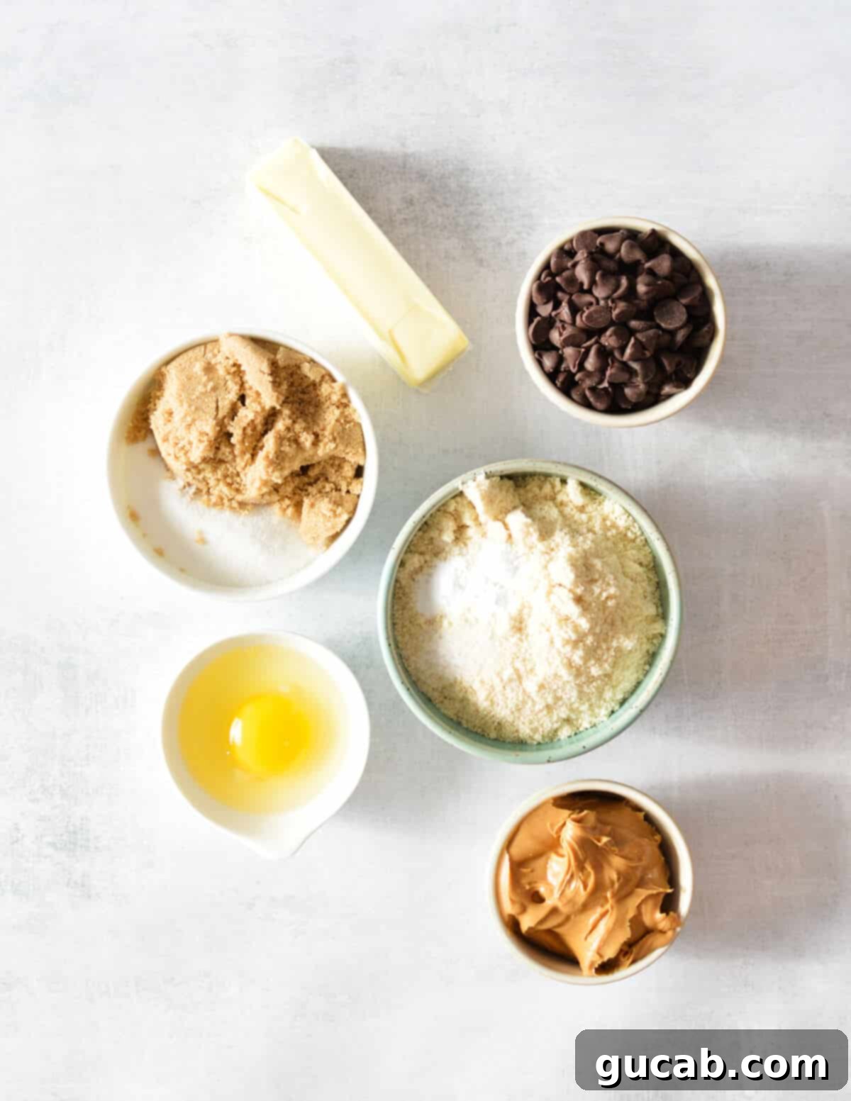 A flat lay of the key ingredients for almond flour peanut butter cookies: butter, chocolate chips, white sugar, brown sugar, almond flour, egg, and a jar of creamy peanut butter.