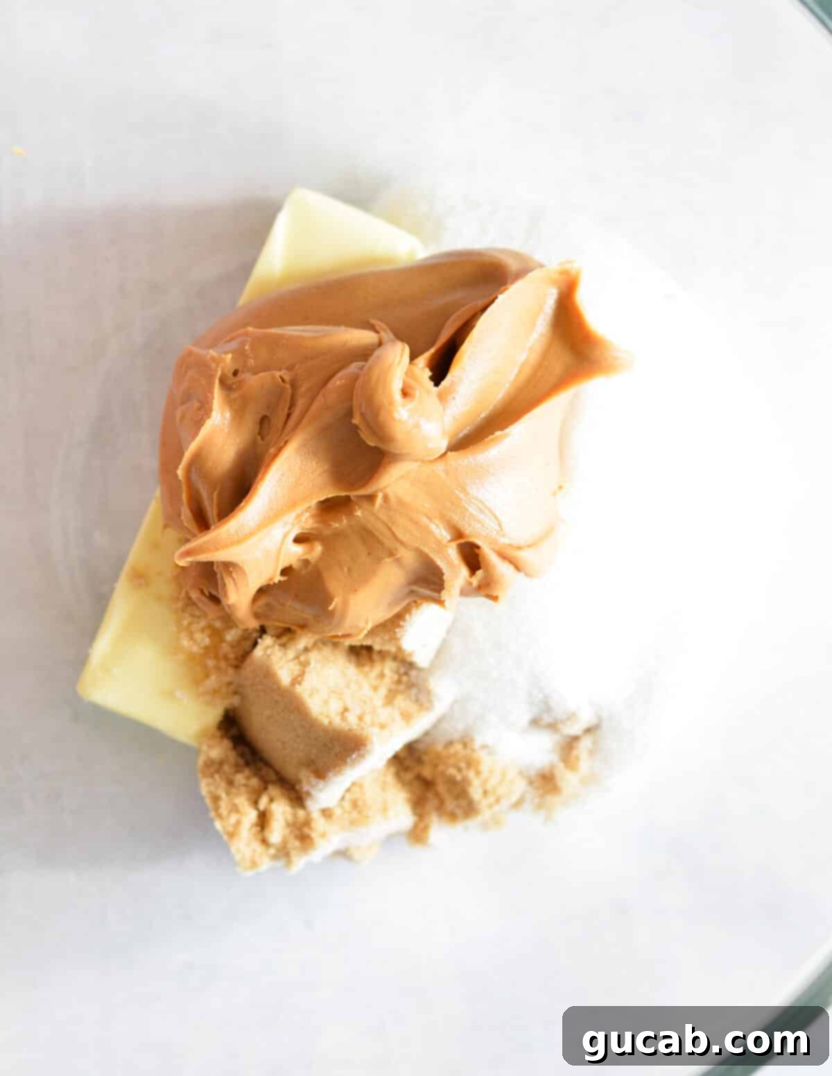 A bowl containing creamy peanut butter, granulated sugar, brown sugar, and softened butter before mixing.