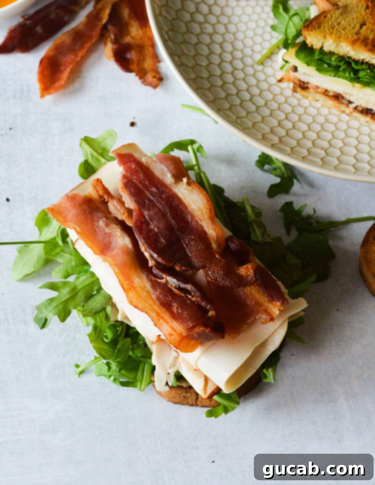 Building a turkey sandwich: toasted sourdough with arugula, slices of deli meat, cheese, and crispy bacon layered on top.