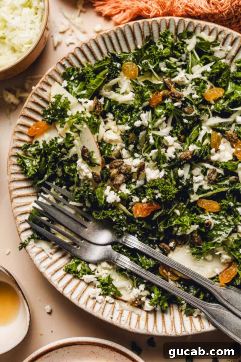 A beautifully plated fall kale salad featuring vibrant green kale, red apple slices, white feta cheese crumbles, golden raisins, and golden-brown maple pepitas, all drizzled with dressing.