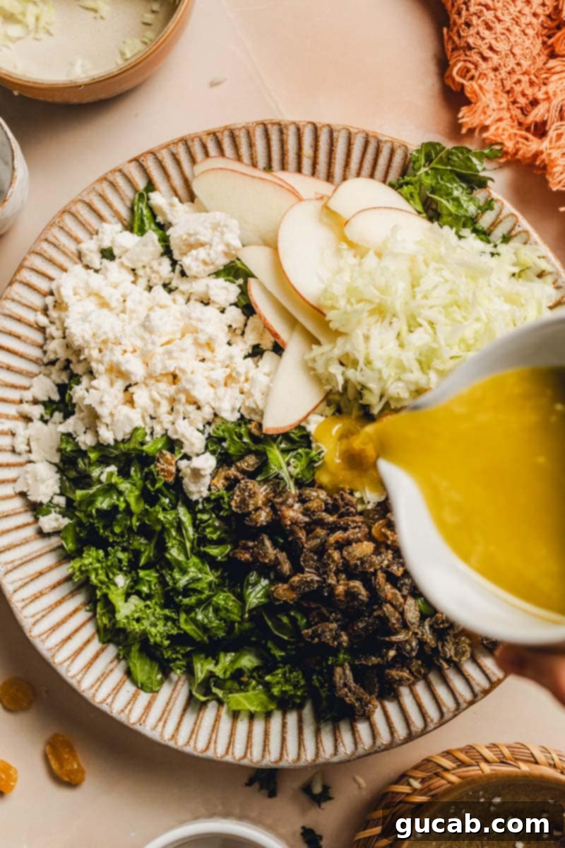 A stream of homemade maple vinaigrette being drizzled generously over the assembled kale salad, which contains apples, pepitas, cabbage, and feta cheese.