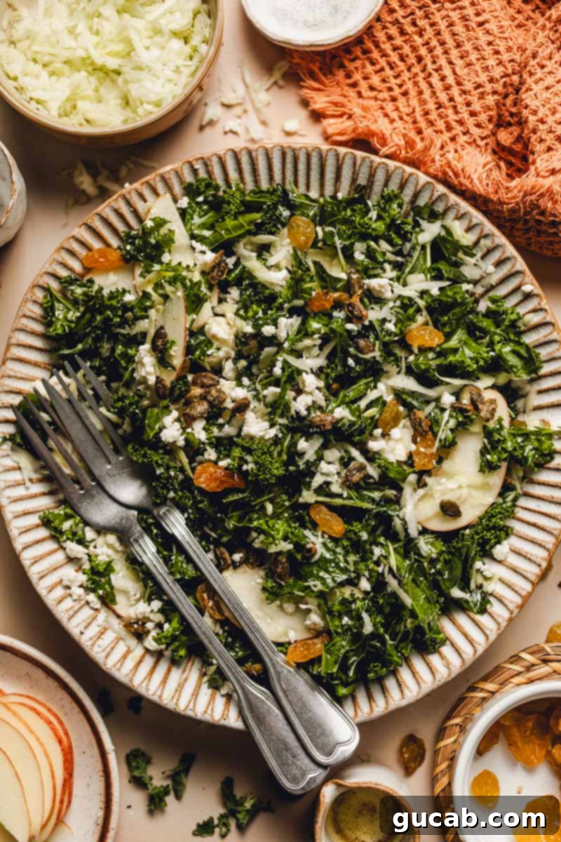 A serving of the fall kale salad, elegantly arranged on a white plate, showcasing the diverse textures and colors of kale, cabbage, golden raisins, apple slices, feta, and maple pepitas.