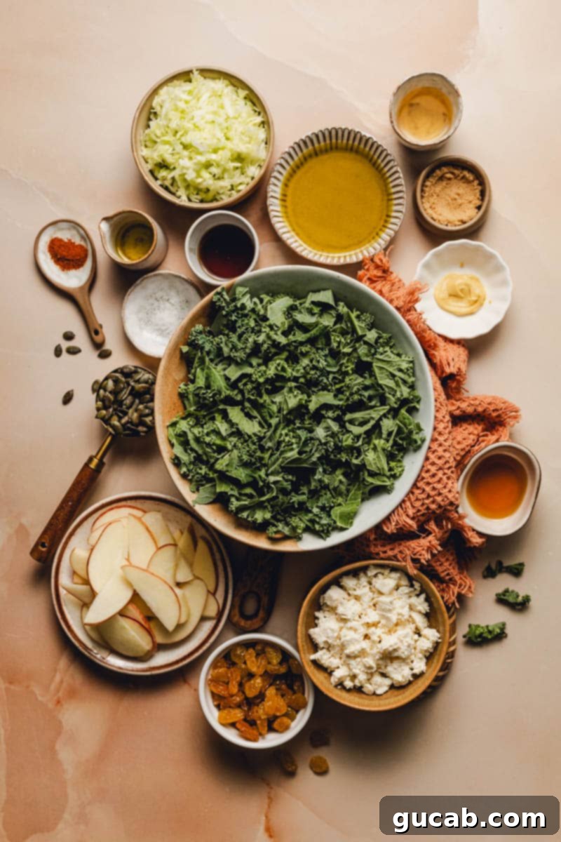 A spread of key ingredients for fall kale salad: fresh kale, sliced cabbage, a bottle of maple syrup, pepitas, bright red apple slices, crumbled feta cheese, golden raisins, olive oil, apple cider vinegar, and various spices.