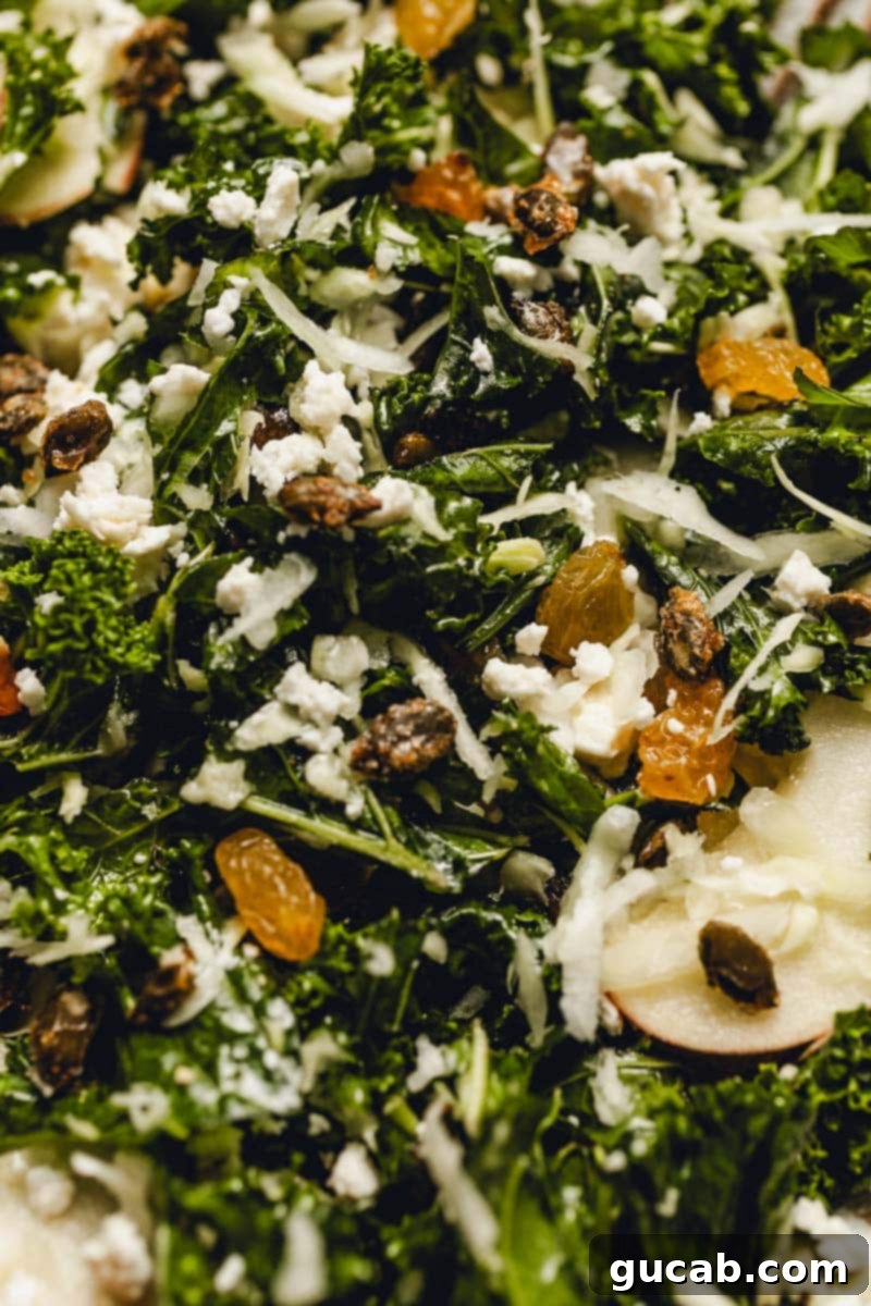 Close-up of the autumn kale salad, showcasing massaged kale, shredded cabbage, glistening golden raisins, vibrant apple slices, and chunky maple pepitas, all coated in a rich dressing.