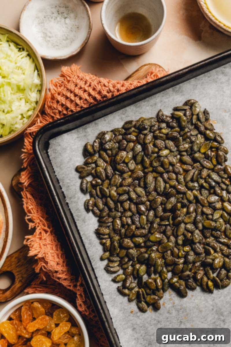 Pepitas coated in maple syrup and chili powder spread evenly on a parchment-lined sheet pan, ready for the oven.