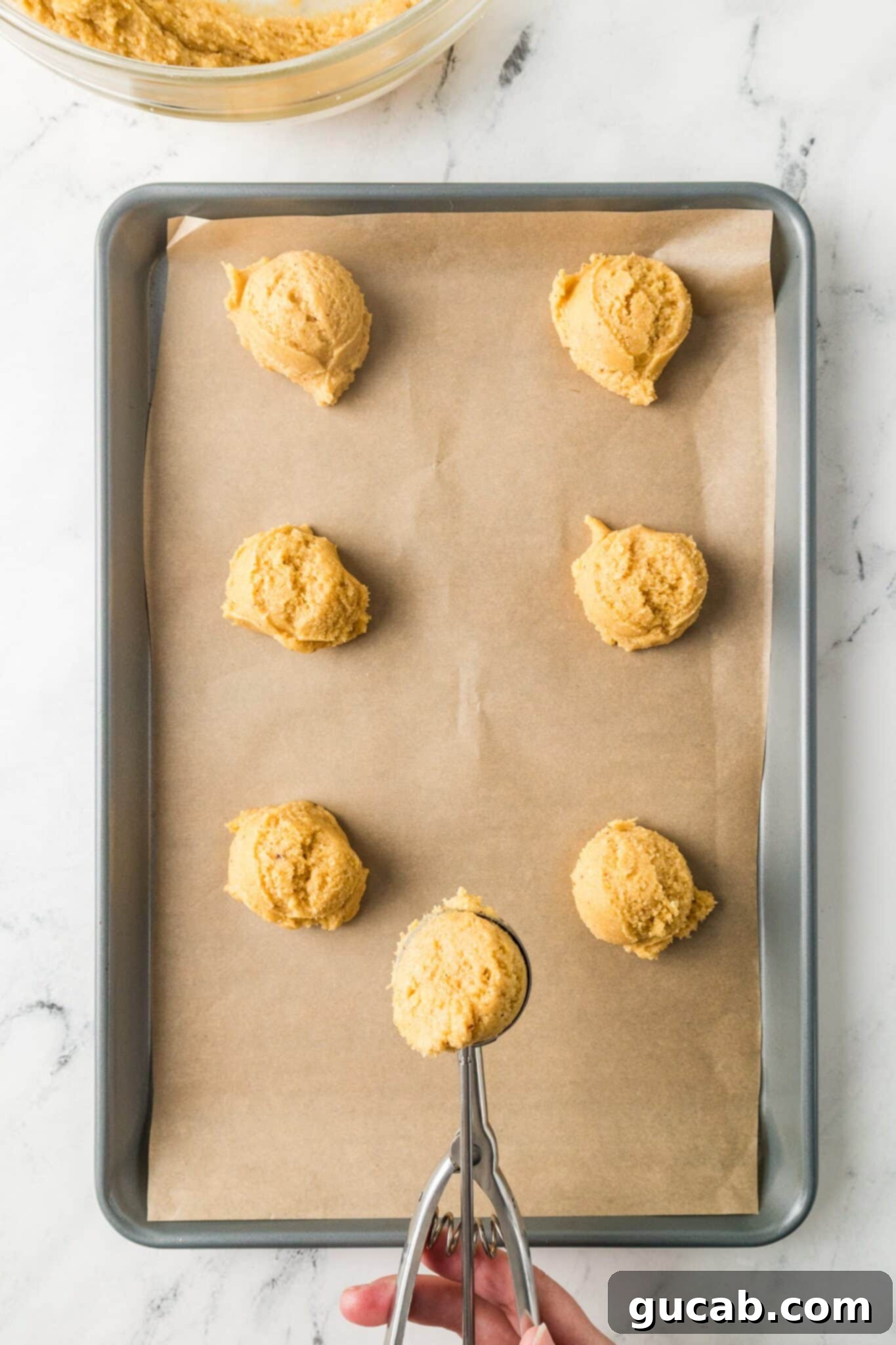 A cookie scoop depositing perfectly rounded balls of cornmeal cookie dough onto a parchment-lined baking sheet.