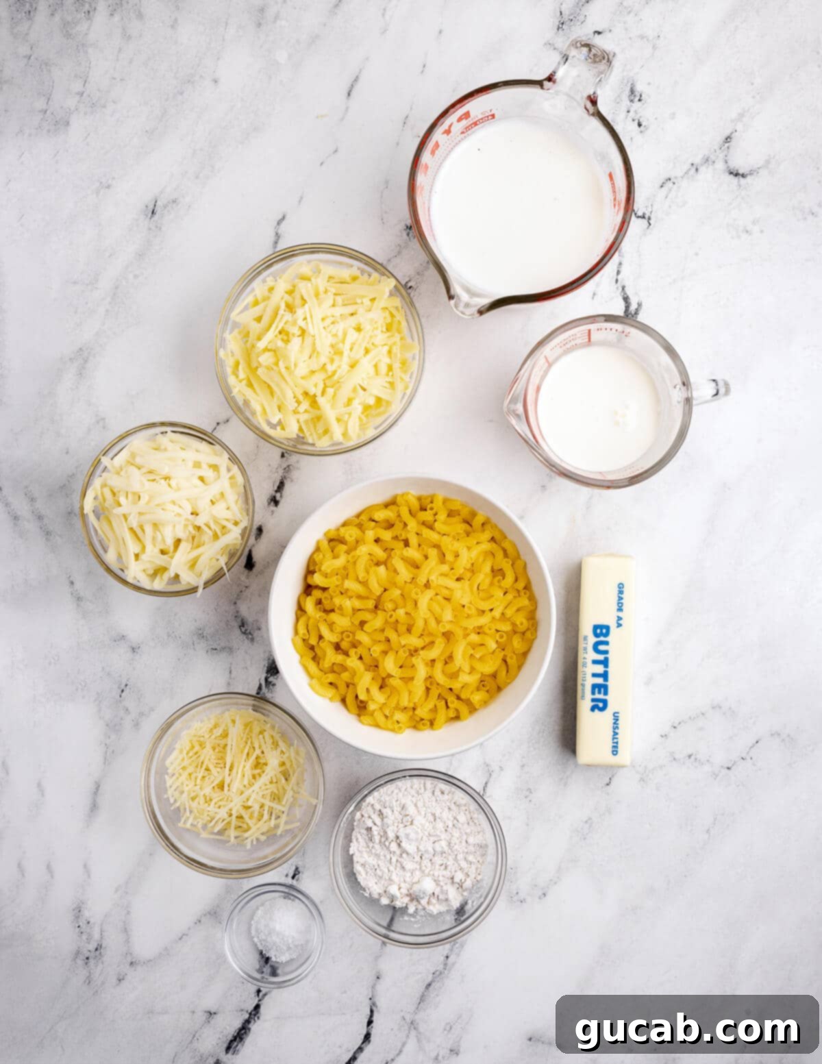 A flat lay of all the essential ingredients for gluten-free mac and cheese: milk, heavy cream, gluten-free pasta, butter, Parmesan, mozzarella, shredded cheddar, and gluten-free flour.