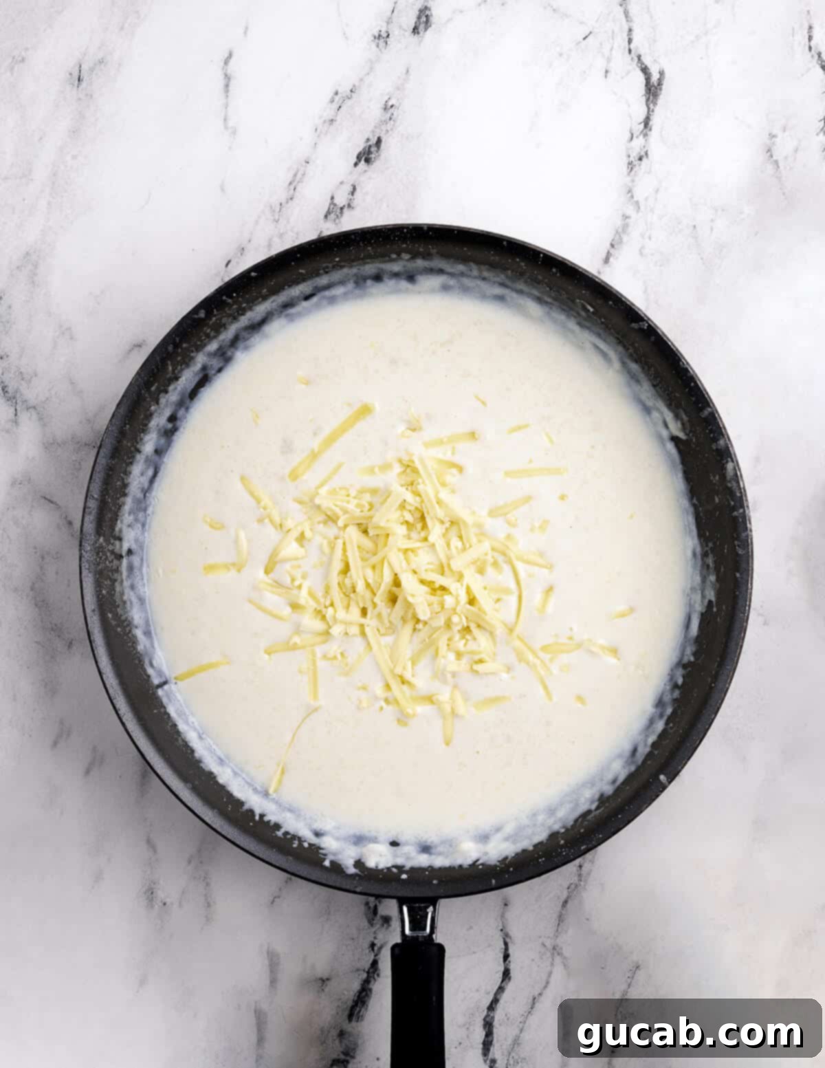 Gradually adding shredded cheese to the creamy milk and roux mixture in a skillet.