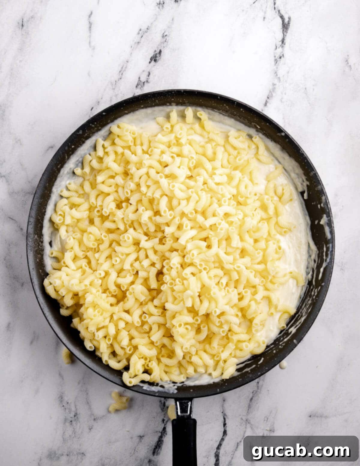 Cooked gluten-free pasta being added to the creamy cheese sauce in a large skillet, ready to be mixed.