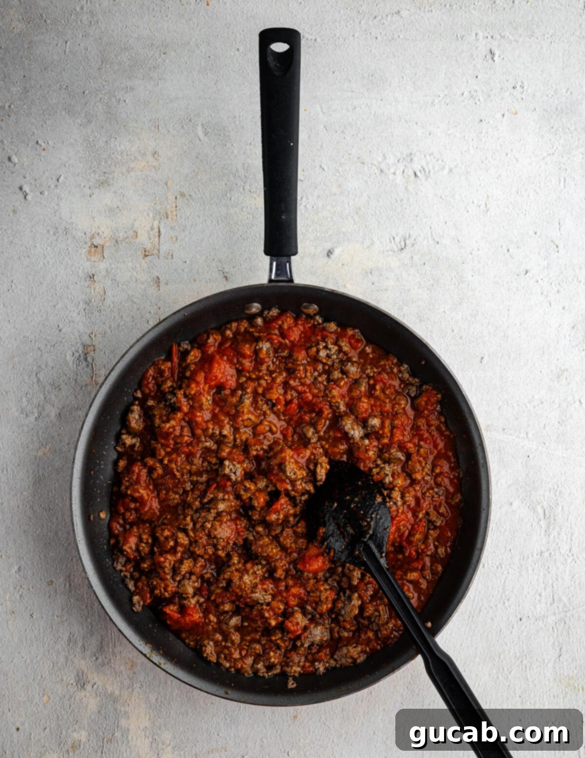 Marinara sauce being stirred into cooked ground beef in a skillet, creating the savory meat sauce.