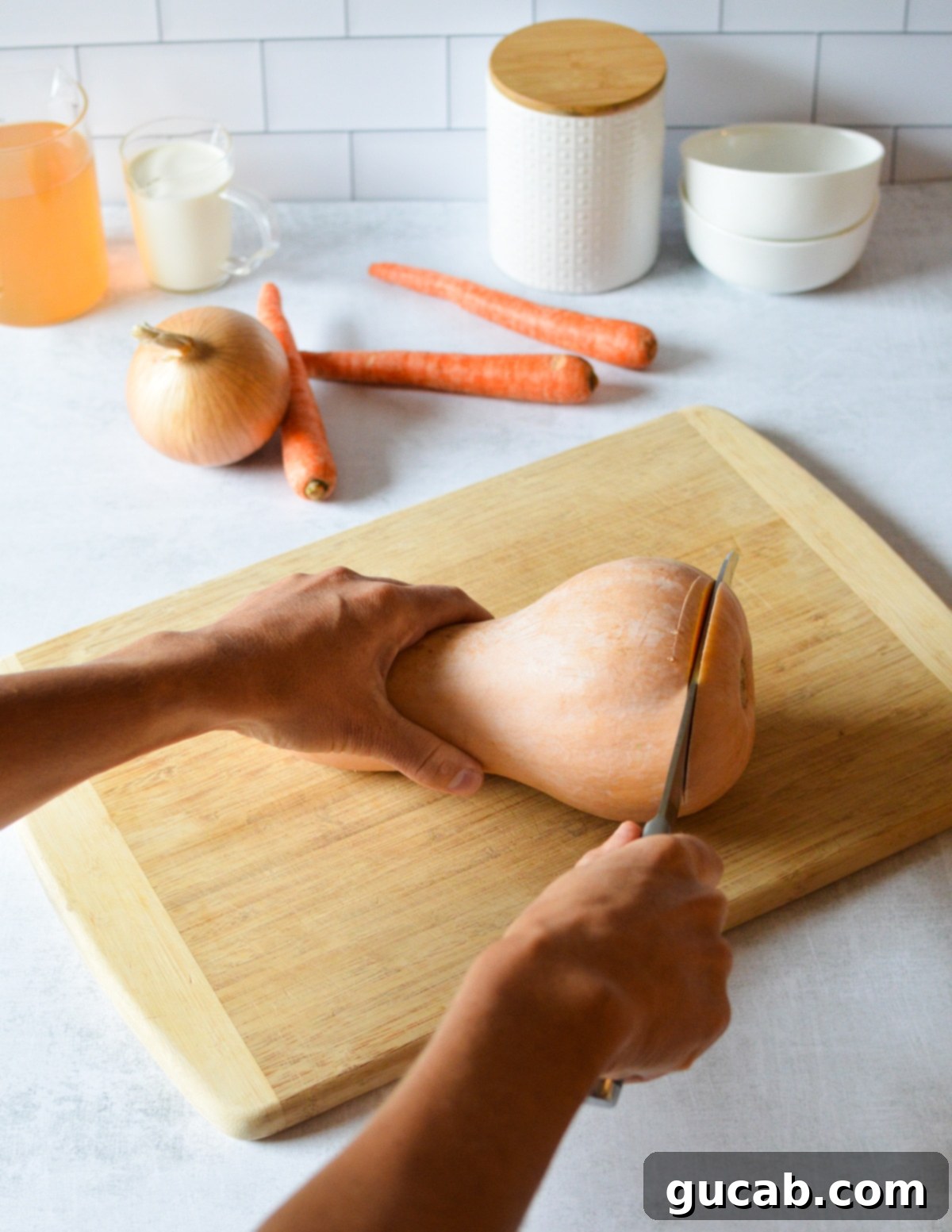 A hand carefully cutting the end off a butternut squash with a chef's knife on a wooden cutting board.
