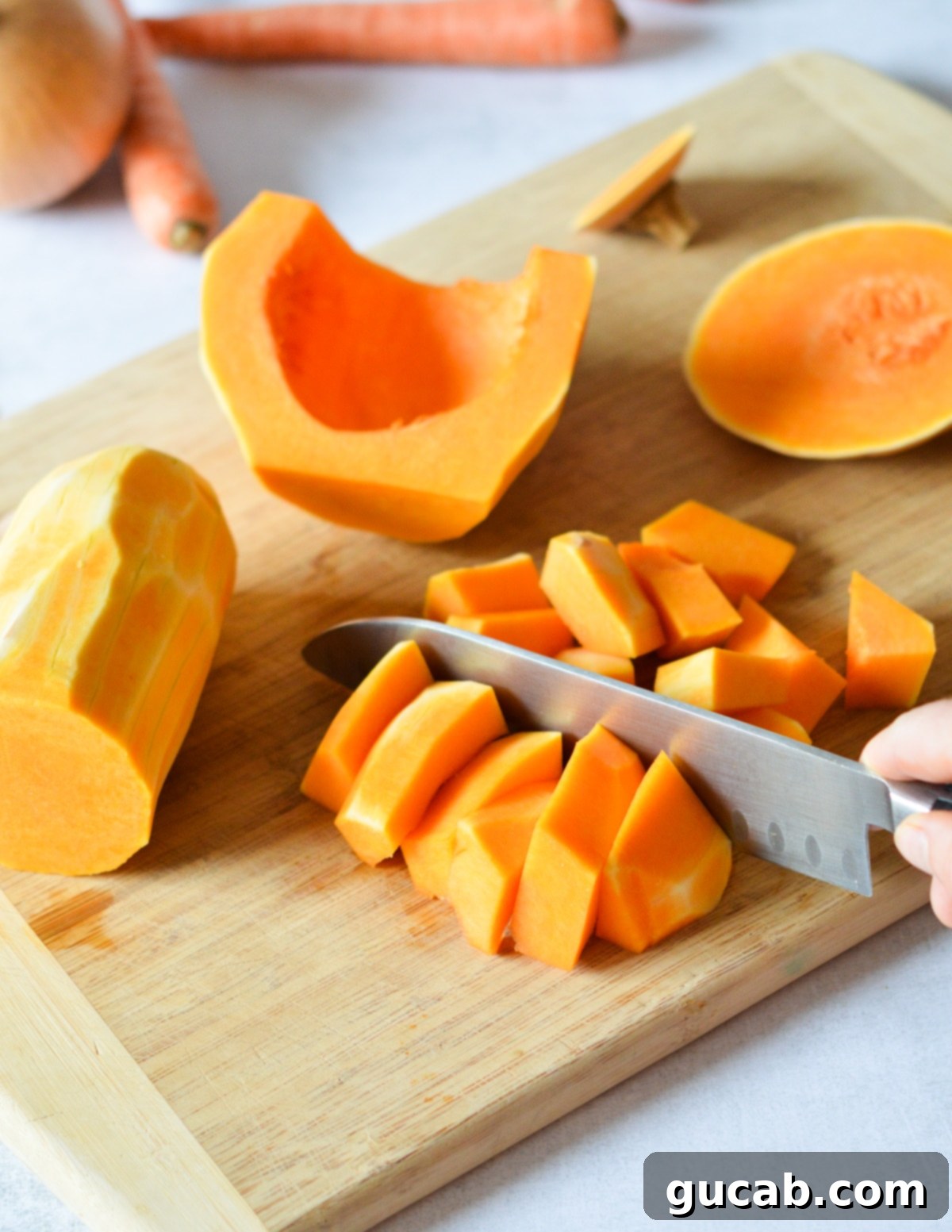 A close-up of a knife cutting butternut squash strips into uniform chunks on a wooden cutting board.