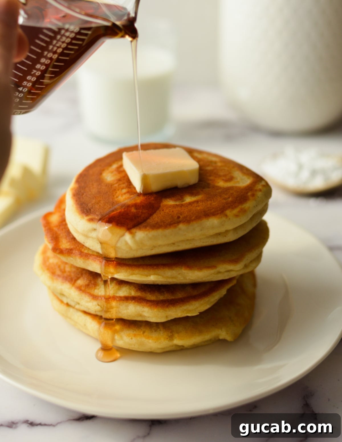 A hand drizzling golden maple syrup over a stack of warm, fluffy gluten-free pancakes.