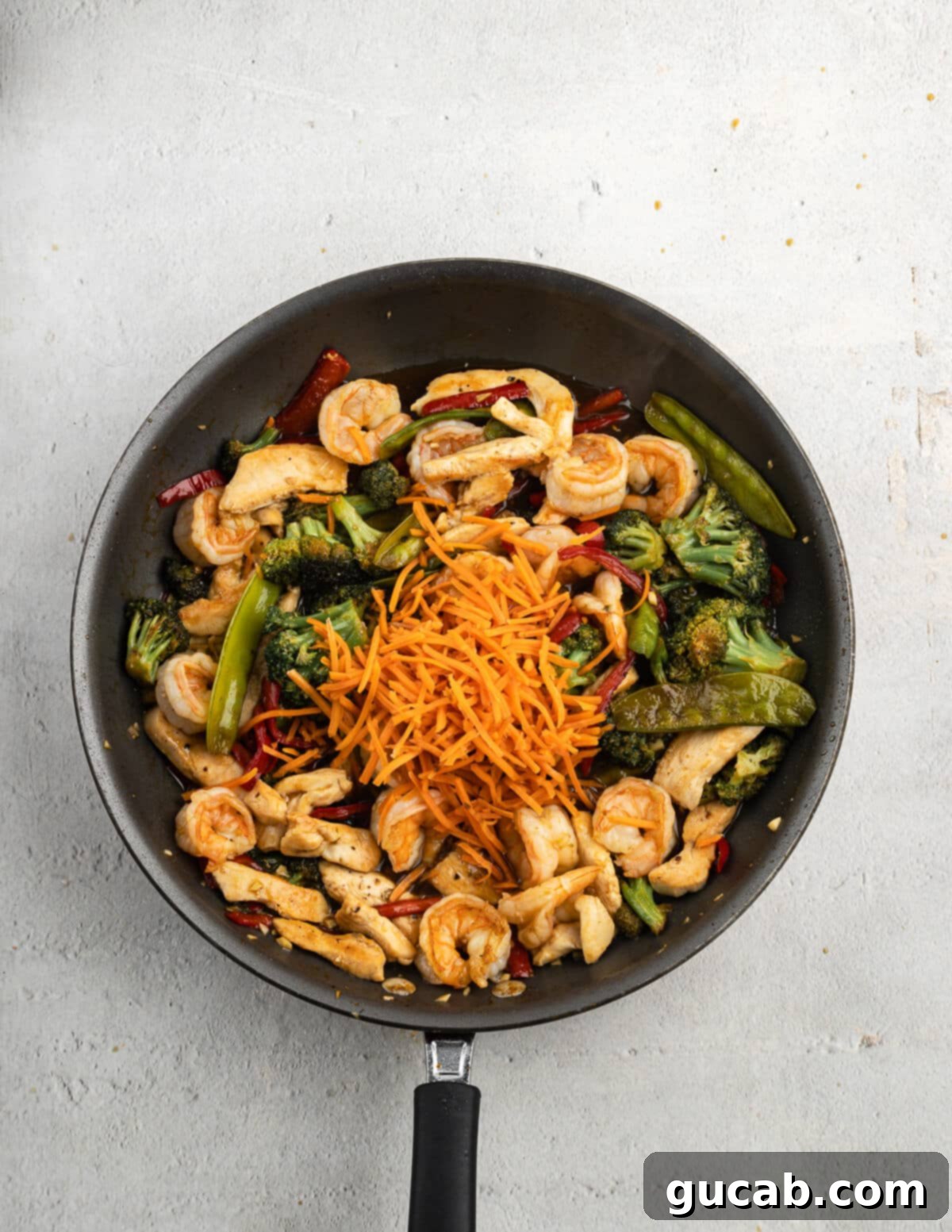 Cooked shrimp and sautéed vegetables in a skillet, topped with fresh matchstick carrots, waiting for the final addition of chicken and sauce.