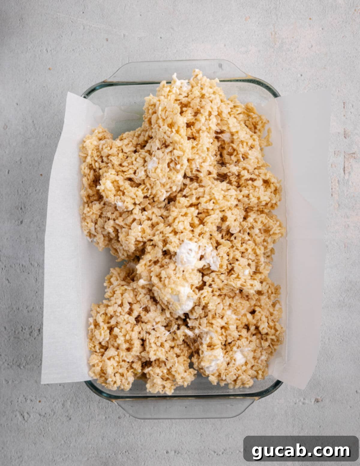 Rice crispy mixture being pressed into a baking dish using a piece of parchment paper.