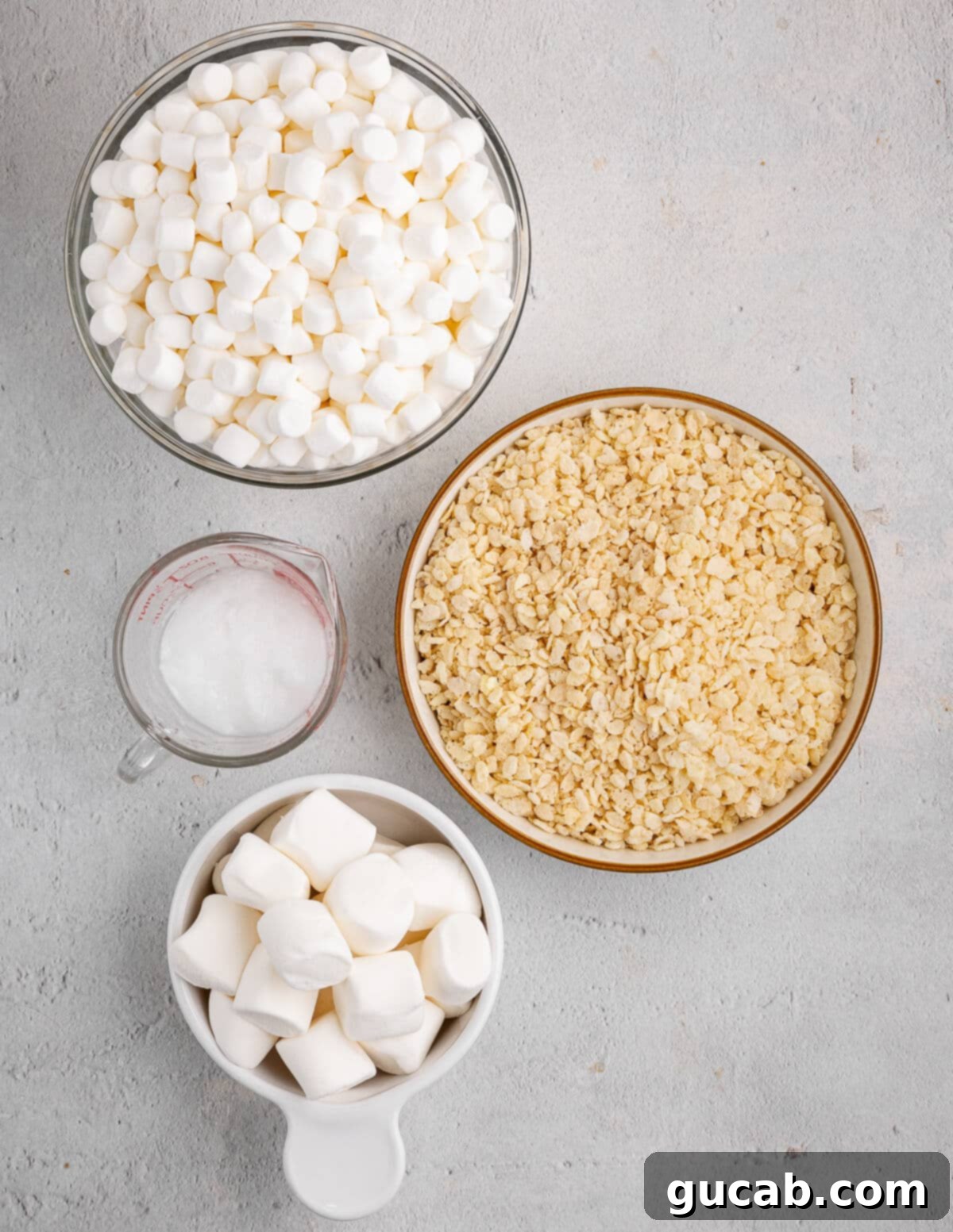 A flat lay photograph showcasing the key ingredients: a bowl of mini marshmallows, a box of rice krispies cereal, a jar of coconut oil, and a pile of large marshmallows.