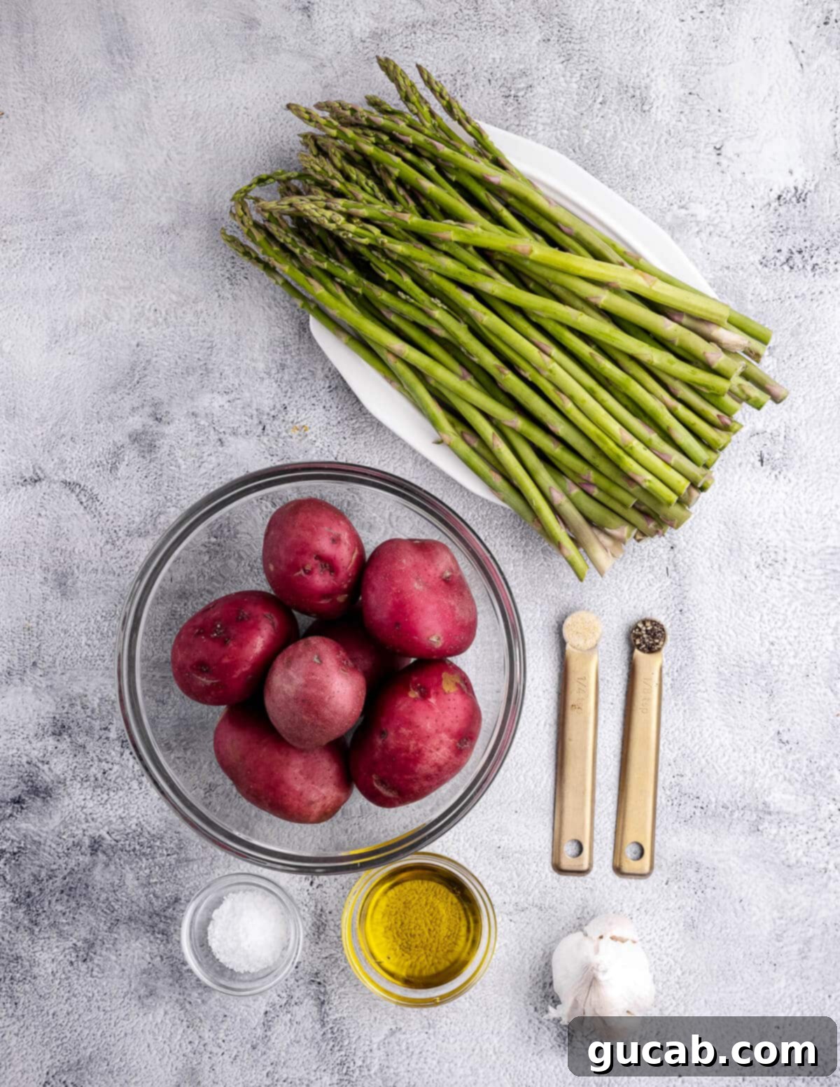 Close-up of the key ingredients laid out: fresh asparagus spears, vibrant red potatoes, whole garlic cloves, a bottle of olive oil, salt, pepper, and onion powder.