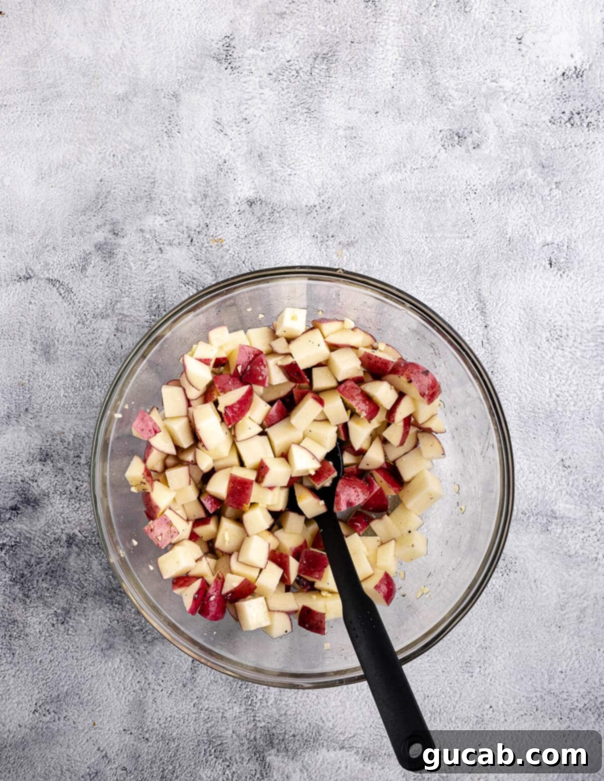 A bowl filled with evenly diced red potatoes, seasoned and ready for roasting, with a spoon resting inside.