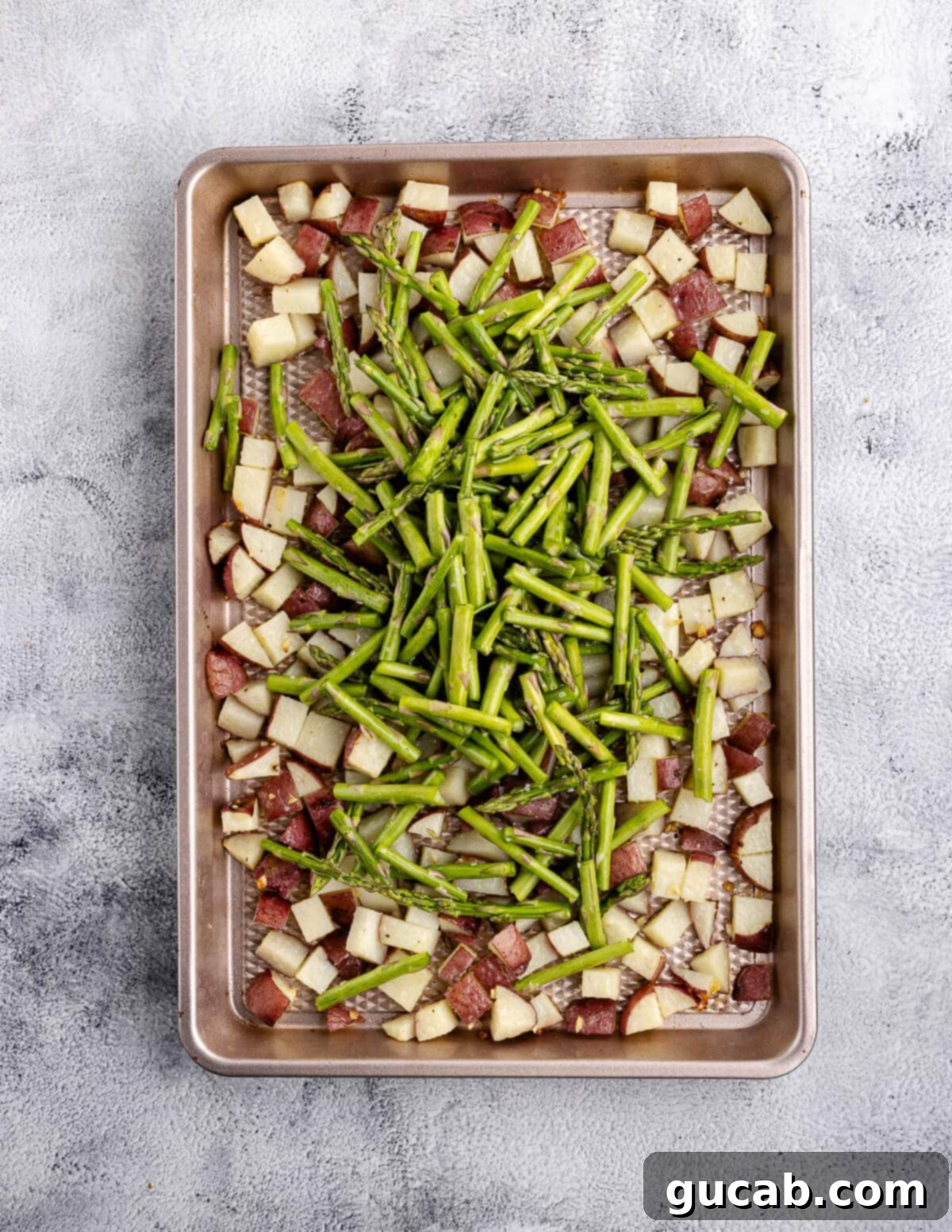 Partially roasted potatoes on a sheet pan, with freshly seasoned cut asparagus spears added on top, ready for tossing.