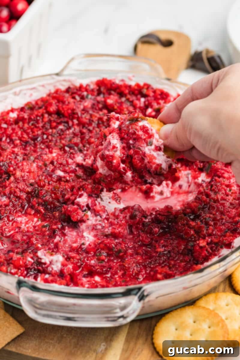 Hand scooping a spoonful of cranberry cream cheese dip onto a Ritz cracker, showcasing its creamy texture and vibrant red topping, ready for a festive holiday gathering.