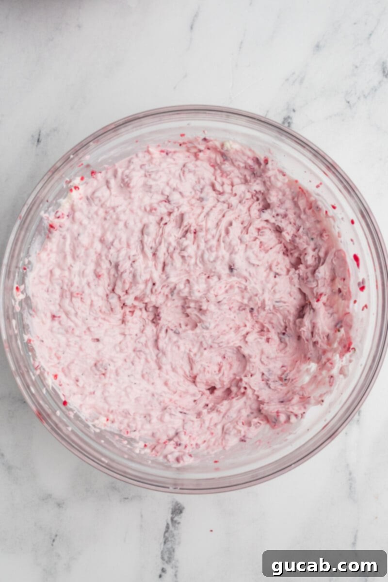 A bowl of cranberry-infused whipped cream cheese mixture, showing a pale pink hue and fluffy texture, ready to be spread as the base of the dip.