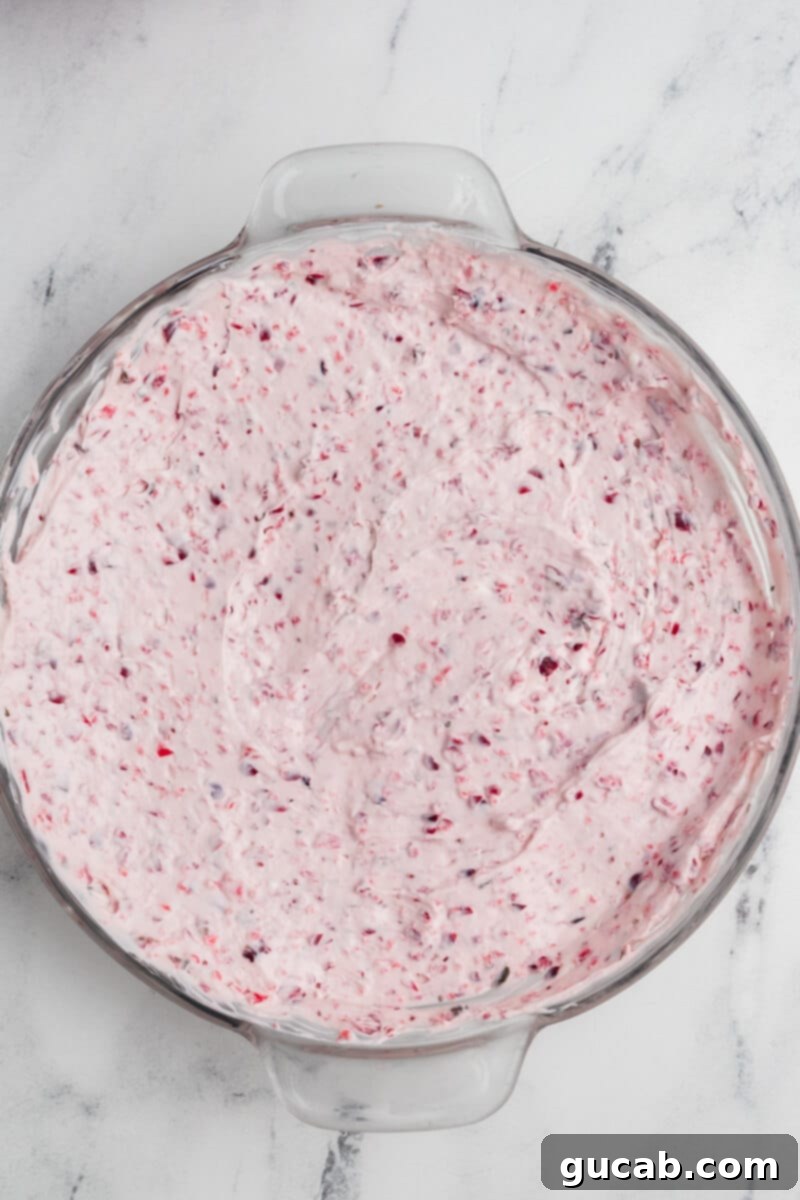 Whipped cranberry cream cheese spread thinly in a white pie dish, forming the base layer for the dip, ready for the chutney topping.