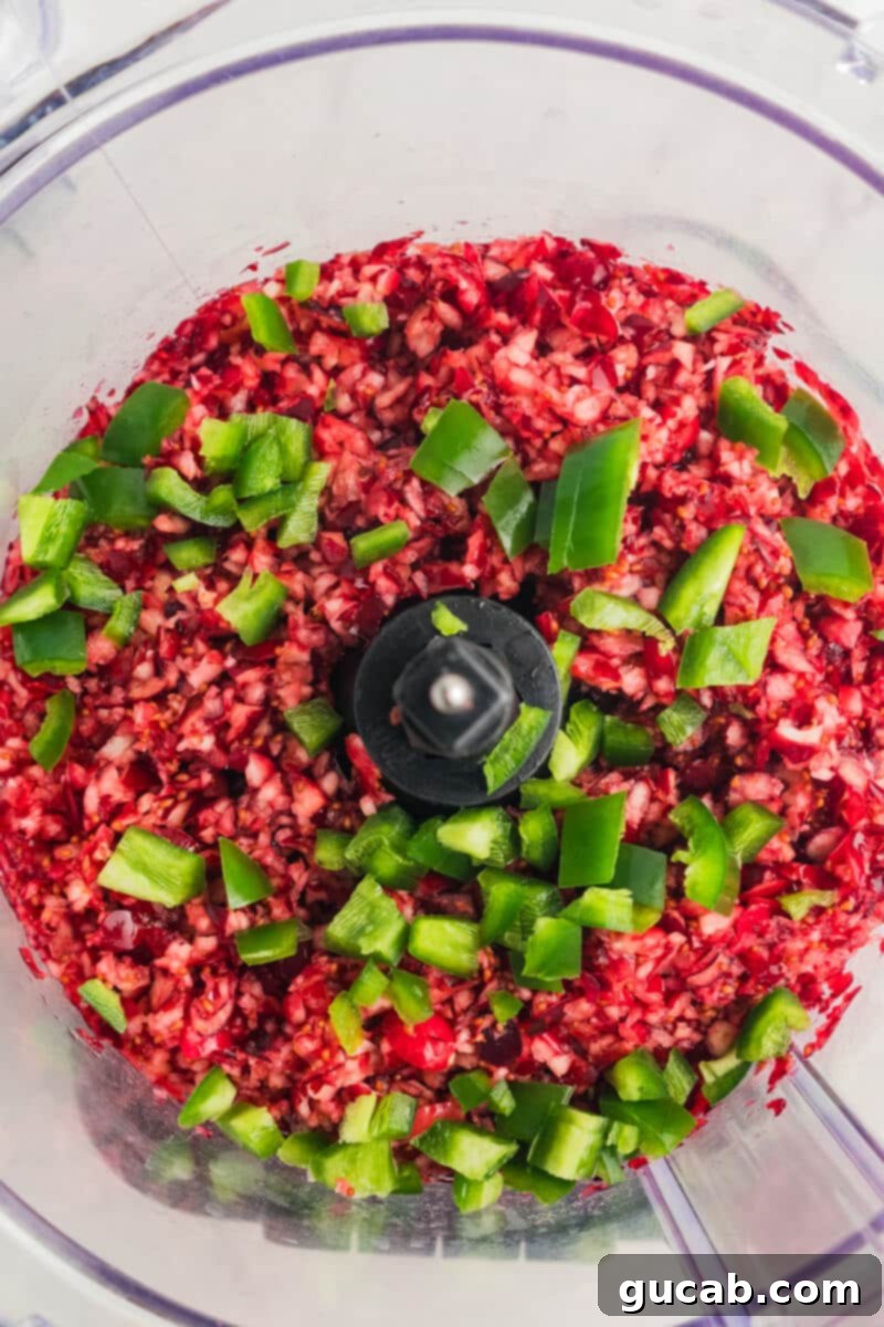 Fresh cranberries and a whole jalapeno placed inside a food processor bowl, ready to be processed for the chutney component of the dip.