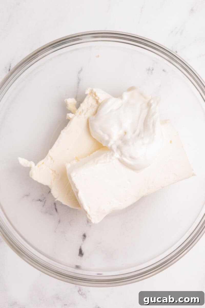 Two blocks of cream cheese and a bowl of sour cream in a mixing bowl, ready to be beaten into a fluffy dip base.
