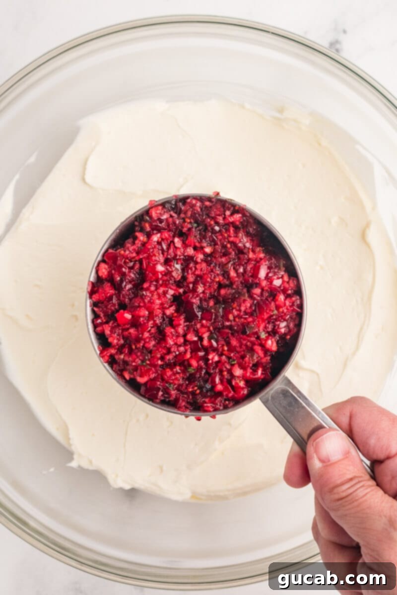 A cupful of vibrant cranberry chutney being spooned over a bowl of fluffy whipped cream cheese, showcasing the optional step of mixing some chutney into the base.