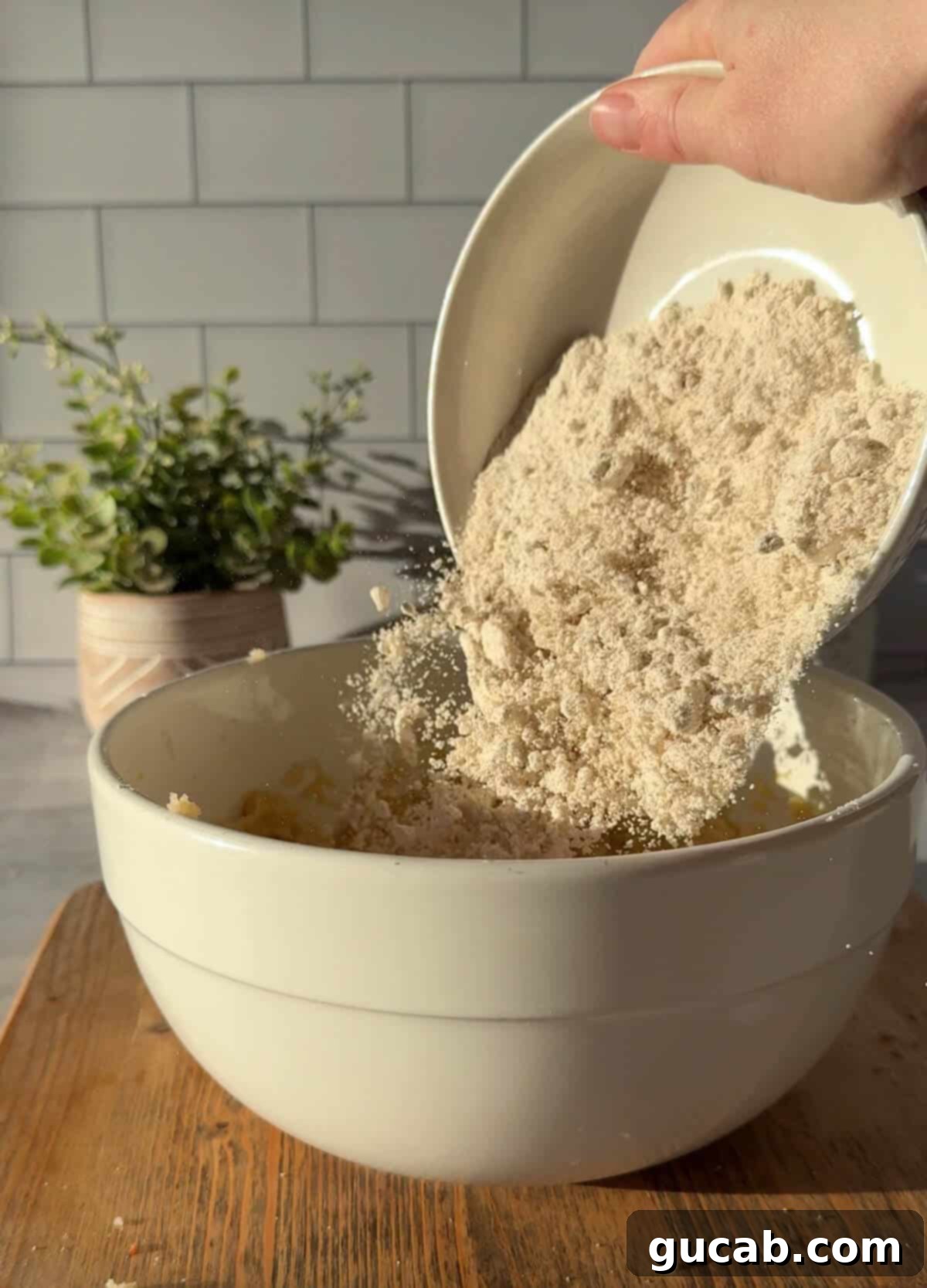 Oat Flour Double Chocolate Chunk Cookies 5 A hand gently pouring oat flour into a bowl of creamed butter, sugar, and eggs, illustrating the addition of the dry ingredients.