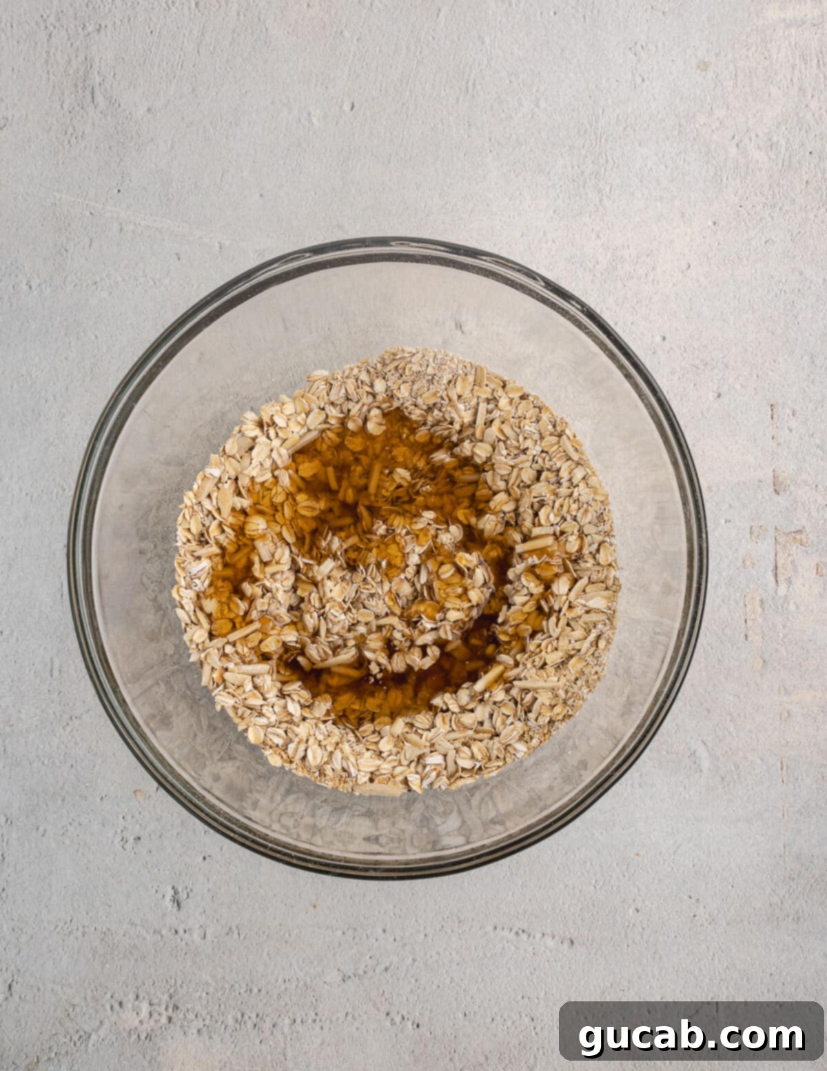 Almond Butter Crunch Granola 7 A bowl of oats with maple syrup and almond butter being mixed together, showing the initial stages of the granola preparation